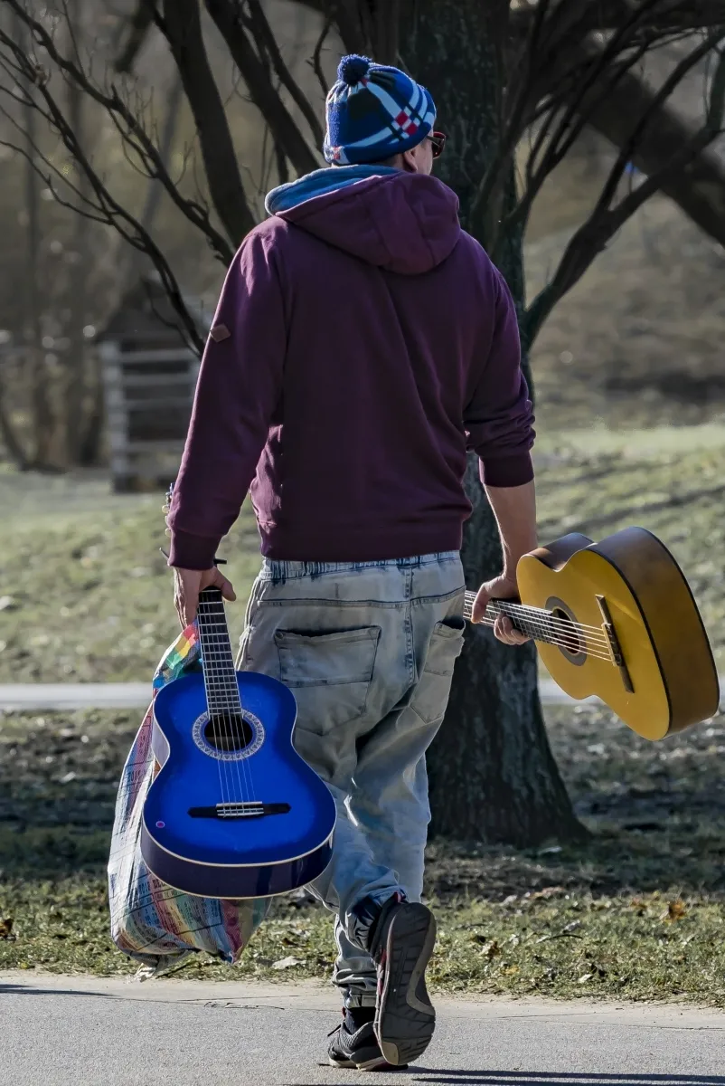 Man walking away through a park carrying a blue guitar and an acoustic guitar